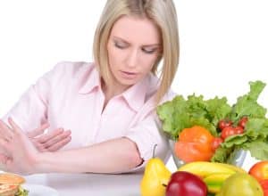 fasting mimicking diet: woman pushing away a hamburger on a plate + rainbow of vegetables on a white counter beside her